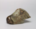 A 3D jade sculpture of a buffalo head that faces the left. The head has carved lines to show horns, ears, eyes, and the snout. The sculpture is pale green in color with spots of dark brown and orange.