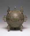 A cast bronze orb that has three legs holding it up, two are visible. The orb is decorated with a geometric, swirling pattern that is mirrored down the vertical and horizontal centers. The legs are made of a circle and a swirled line that goes out, three on the bottom and three on the top. Two small circle handles are on the left and right.