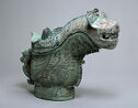A blue-green cast bronze vessel that has a wide, stout foot, a stout body, a large spout that has the head of a wide-mouthed tiger on top, and is covered in fine inscribed swirling lines.