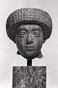 A dark gypsum sculpture of a head that stands on a pole on a pedestal. It has large, almond shaped eyes and a large, rounded headpiece that goes out around the head. The headpiece is thick and has a round texture.