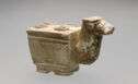 Brownish cube-shaped vessel with four holes at the top and a neck-like handle, terminating in a water buffalo head.