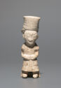 A jade statuette of a short person standing upright. They are wearing a wrapped robe that shows their feet and arms. Their hands are in front of their stomach. Their head is large with carved lines detailing the face. They are wearing a tall, cylindrical 