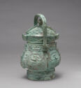 A gray-green bronze vessel with a lid and a middle handle on top of it. It is smaller at the top and wider at the bottom with a thick foot. It is inscribed with detailed, swirling designs all over.