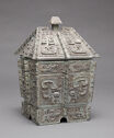 A grey cast bronze box-shaped vessel with a sloped lid. The entire piece is inscribed with detailed, swirling designs with the bottom-center having a goat-like inscription.