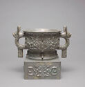 A mottled grey cast bronze cup with a thick, cube-shaped bottom and two decorated large, semi-circle handles. The handles have a protruding form on top and bottom of them. The outside of the cup is decorated with a swirling pattern of thick and fine lines.