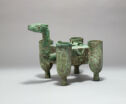 A blue-green cast bronze four-legged container with an upright dragon neck and head. The four corners of the object are in the shape of narrow cups and the center is a solid square. It is inscribed with a pattern throughout.