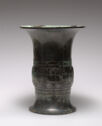 A mottled black and green cast bronze vessel that stands on a wide foot, has a slightly round body, and a tall, flared out neck. The body is decorated with two horizontal bands of engraved patterns.