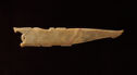 A jade sculpture that is long in shape with the left side being wide with rounded engraved details and the right side coming to a narrow, blunt edge. It is colored golden yellow. The sculpture has a crack diagonally down the middle with red coloring around it.