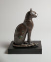 A bronze sculpture of a seated cat on a black plinthe. The cat sits upright with its tail around its front paws and ears up.
