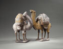 Two figurines of camels standing and facing each other. One is mottled grey and the other is yellow-tan. Each has stacks of rolls and flat fabrics tied to their backs and small, dark plinthes under their feet.