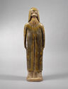 A led-glazed ware figure of a man standing upright and wearing a long, wrapped jacket. He wears a flat headpiece that covers the sides of his head. He has a long, straight beard.