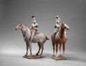 Two earthenware figurines of a man and woman riding horses. The man wears a tall hat and the woman hears her hair on top of her head. Both are wearing a wrapped jacket and pants. Each horse has their four hooves connected by a small plinthe.