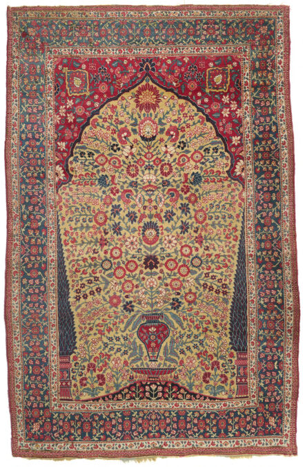 A rectangular carpet with detailed floral design with a patterned green border, a yellow pentagonal shape in the middle that points up, and a small red vase at the bottom. The entire rug is detailed with the floral designs.