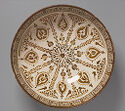 A circular lusterware bowl that is colored white with painted brown details. There is a thick, patterned band along the top edge, large tear-drop shaped designs around the middle, and a floral shaped motif at the center. The floral motif has many waving lines that reach the top edge.