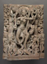 A relief sculpture made of light red sandstone that shows a tall person at the center, standing on a smaller person and surrounded by other small figures. The tall person is holding small objects in their hands and is wearing beaded jewelry.