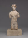 A white-slipped terracotta sculpture of a nude woman with no arms or legs. She stands on a stone block.