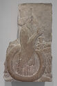 A limestone relief depicting the profile of a standing man wearing a draped robe and a tall headpiece. He holds an object in one hand and his bottom half has a large circle around it.