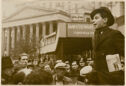 A black and white photograph of man speaking to a crowd in an urban landscape.