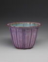 A bright purple ceramic flower pot with vertical lines widely spaced out on the side. The inside is aqua colored. The flat, thin top rim protrudes out. The rim curves to mimic large petals.