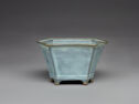 A light blue stoneware vessel with tall flat walls, a flat jutted out lip in the shape of a hexagon, six small feet, and protruding corners along the outside. The top lip makes a hexagonal shape.
