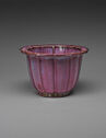 A magenta ceramic flower pot with vertical lines widely spaced out on the side. The flat, thin top rim protrudes out. The rim curves to mimic large petals.