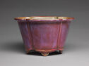 A magenta stoneware vessel with high walls, a flat jutted out lip, and indented lines along the outside. The small feet are cut-out in a slightly swirling shape.