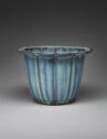 A blue ceramic flower pot with vertical lines widely spaced out on the side. The glaze is a mixture of light blue and a medium, dark blue. The flat, thin top rim protrudes out. The rim curves to mimic large petals.