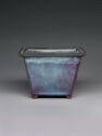 A square-shaped ceramic vessel with tall, straight walls. The top lip is flat and the feet are very small. It is colored bright blue with some purple coloring along the wall edges.