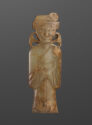 A flat jade pendant of a person that is dark green in color and has been carved with fine lines to outline a wrapped robe, belt, draped sleeves, their face, hair, and a small head piece. There are two protruding, round shaped coming out from the neck with