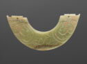 A jade piece that is shaped into a semicircle with the center cut out. It is green in color. There are swirling carved lines throughout the piece that make a pattern. The left and right edges of the piece are horizontal and irregularly-cut with a small ho