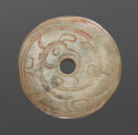 An off-white jade disk with a circle cut out in the middle. There are carved lines that create a broad, swirling pattern throughout the piece. There is some red discoloration.