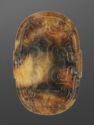A jade plaque that is oval in shape and brown and black in color. There is an engraved symmetrical, swirling pattern at the center of the plaque. There are small holes at the top and bottom corners and sides of the plaque.