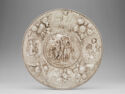 A circular silver dish is decorated with scenes in the center and around the edges.