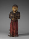 A wooden sculpture of a young boy standing upright. The boy is wearing a long, red piece of clothing that goes from his hips and covers his feet. His hands are together in front of his chest. The boy is bald and is looking down at his hands.