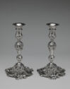 
A pair of tall ornate silver candlesticks with swirl and wave patterned stems and bases stand next to each other.