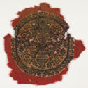 A circular textile fragment with irregular borders depicting flowers in a vase in purple on a yellowed white ground with a red border. 