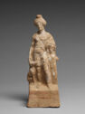 A terracotta statue of a woman standing on a block. A small person is by her left side and a dog on her right. She is wearing a short skirt and straps cross her chest.