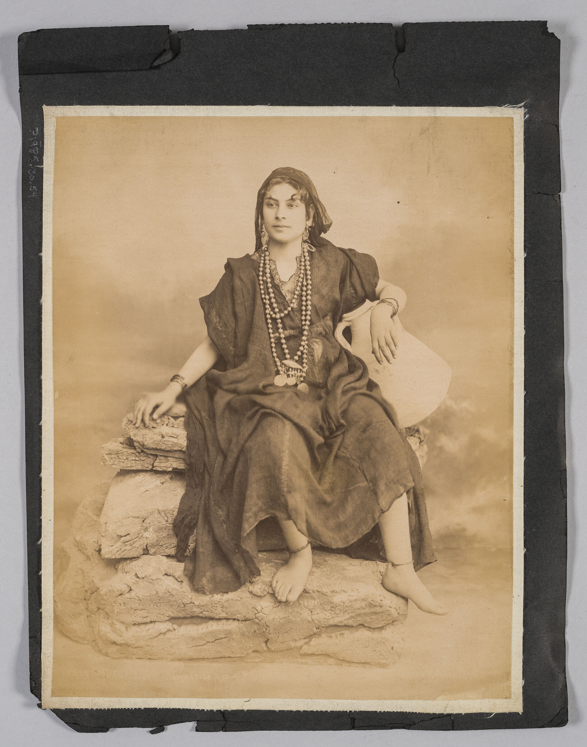 Untitled (Woman In Traditional Dress Seated On Pile Of Stones, Holding A Jug Under Her Left Arm)