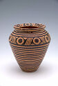This ceramic pot has a tapered bottom and rounded shoulder leading to a flared opening on top. It is decorated with horizontal lines with bullseye designs on the shoulder and inner lip.