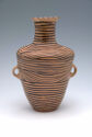 This is a light brown, circular ceramic vase with two circular handles below the shoulder and a long spout with a flared opening. It has dark brown horizontal lines as decoration.