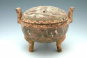 An earthenware vessel that is round in body and stands on three narrow legs. It has a round, flat lid and two handles that stand straight up on the left and right. The body of the piece is colored green with a red and white swirling pattern. The handles and legs are brown.