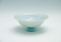 A blue-green porcelain cup that stands on a small, rounded foot. The bowl is low and wide and flares slightly at the lip. There are small indents around on the lip.