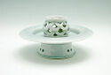 A pale blue-green porcelain piece that stands on a wide, round hollow foot. The body juts out like a flat plate in the center. On top in the center is an openwork round form with a flat top. The openwork piece is made of thick, rounded lines that make a pattern.