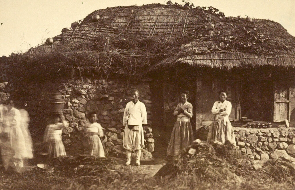 A Country Family Dressed In Traditional Robes And Standing Before A Farm House With Thatched Roof