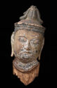 A painted wooden sculpture of a head that is facing the viewer. The head is tilted to the right and is round in shape. The head has long earlobes and wears a black headdress.