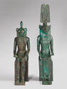 Two bronze figures with lion heads and human bodies sit next to each other. The one on the left wears a skirt and a cobra on its head. The one on the right wears a long dress and a tall headpiece.