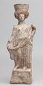 A terracotta statue of a woman standing on a stone block. She wears a long, draped dress and her hair is braided on top of her head. A large object sits on top of her head.  