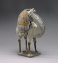 A camel figurine standing and facing the right of the viewer. It’s neck is bent back with its mouth open. It is colored off-white and grey. It is carrying a stack of rolled and flat goods tied to its back. There is a small, dark plinthe under all of its feet.