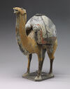 A camel figurine standing and facing the left of the viewer. It is colored yellow-tan. It is carrying a stack of rolled and flat goods tied to its back. There is a small, dark plinthe under all of its feet.