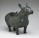 A green-black cast bronze vessel that is in the shape of a standing four-legged, hooved animal. The creature’s body is thick and round with a long, downturned tail. Its neck is thick and its head has a long, small snout, round eyes, and almond-shaped ears that point straight up. There is a curved form in the center of its back.